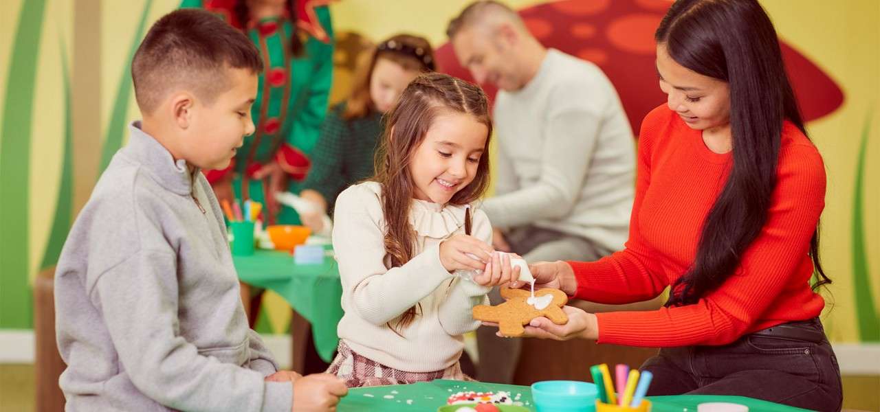 Girl squeezes white icing onto a gingerbread cookie held by a woman, while a boy watches; others craft at green-covered tables in a colorful, festive classroom.