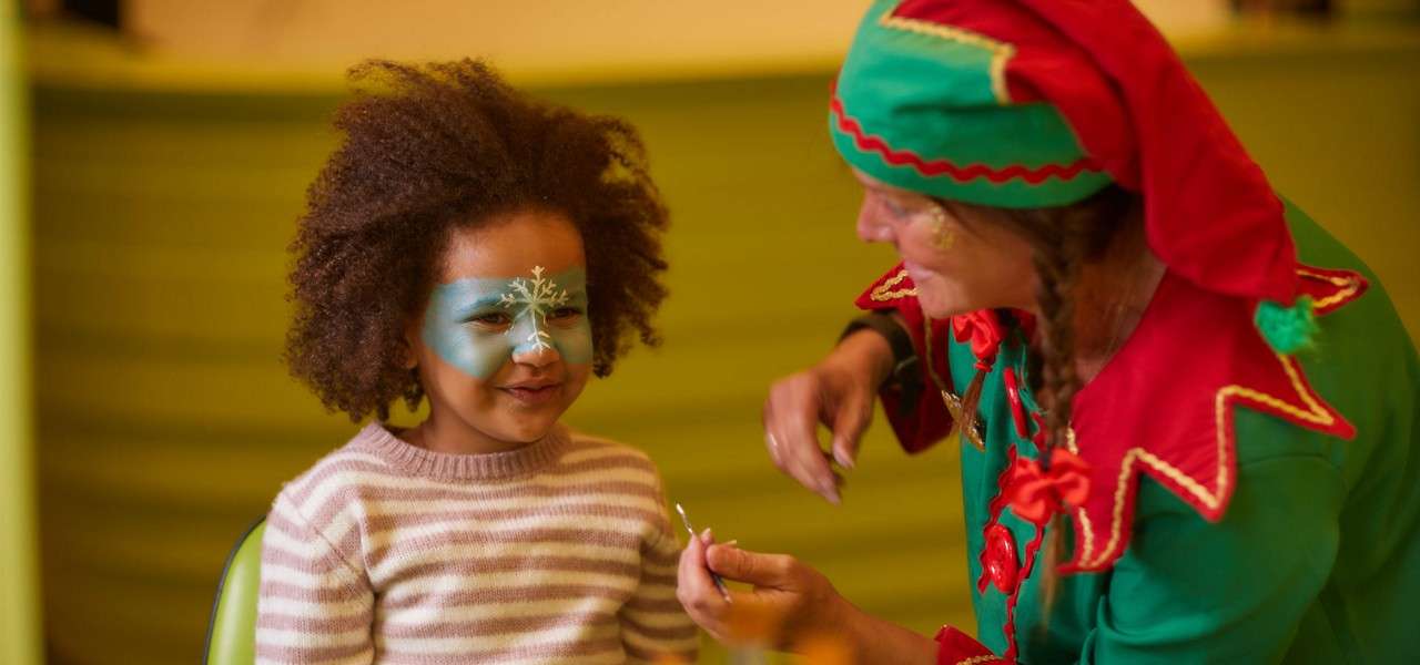 Child smiles while receiving face paint from a costumed performer in green and red holding a brush; indoors with soft lighting, green backdrop, striped sweater, and blue snowflake face design.