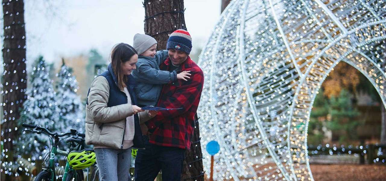 Family consults a map as a parent holds a bundled child, standing beside bicycles amid a festive outdoor display—twinkling light arch, tree trunks wrapped in lights, and snow-dusted evergreens.