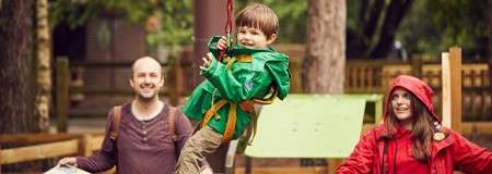 Child in green raincoat rides a low zipline handle, smiling midair. Two adults stand nearby, one holding the line. Wooden fences, trees, and playground structures form a forest adventure-park setting.