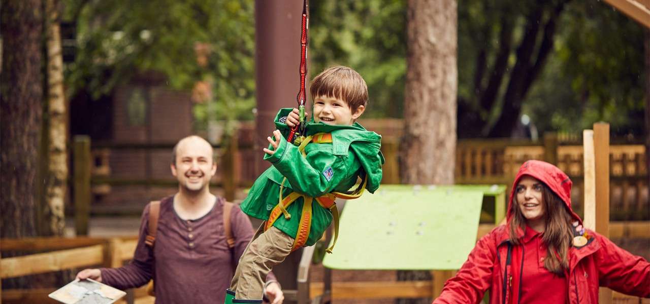 Child in a green raincoat rides a harnessed zipline, smiling; two adults, including one in a red hooded jacket, watch in an outdoor wooden playground with trees in the background.