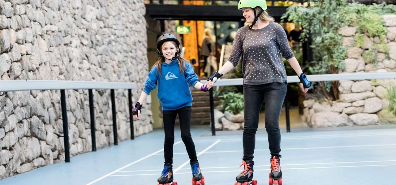 Two skaters hold hands and glide forward, wearing helmets and pads, on a smooth indoor rink bordered by stone walls and railings, with a doorway and plants in the background.