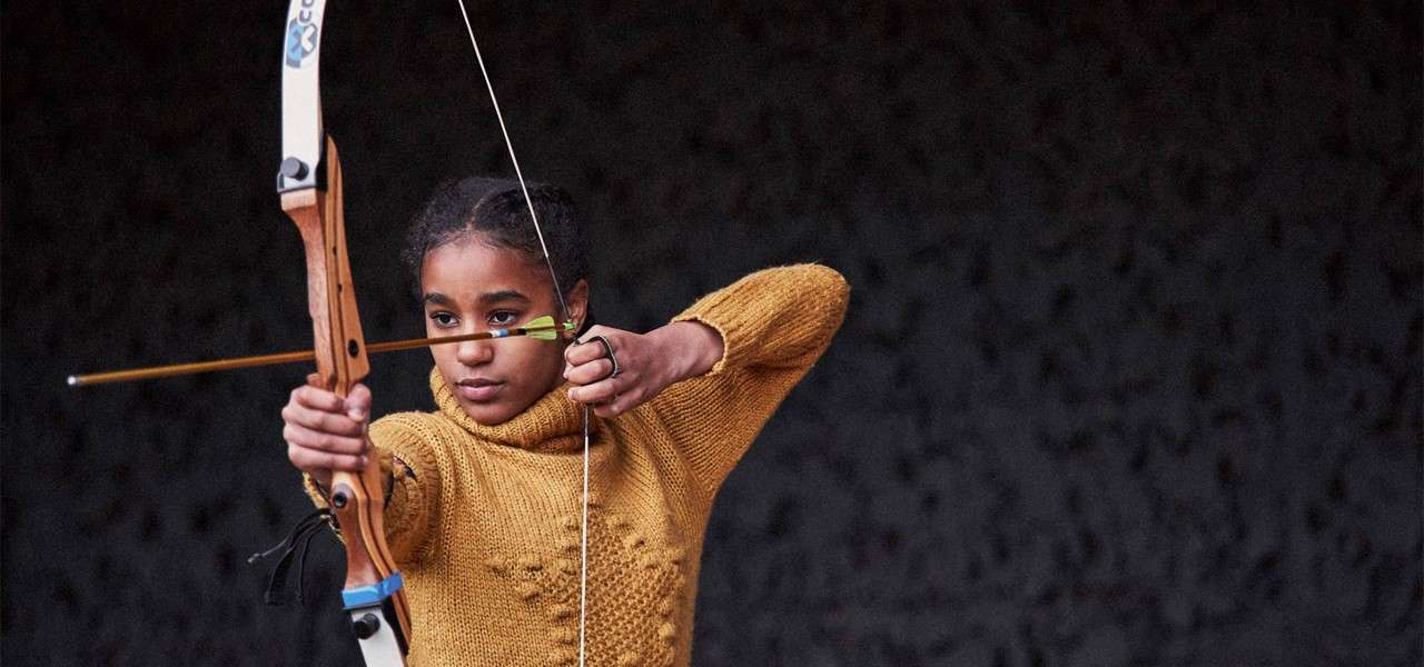 Archer draws a recurve bow, aiming an arrow at target, wearing a mustard sweater, against a dark textured background. Text: “CO” on the bow limb.