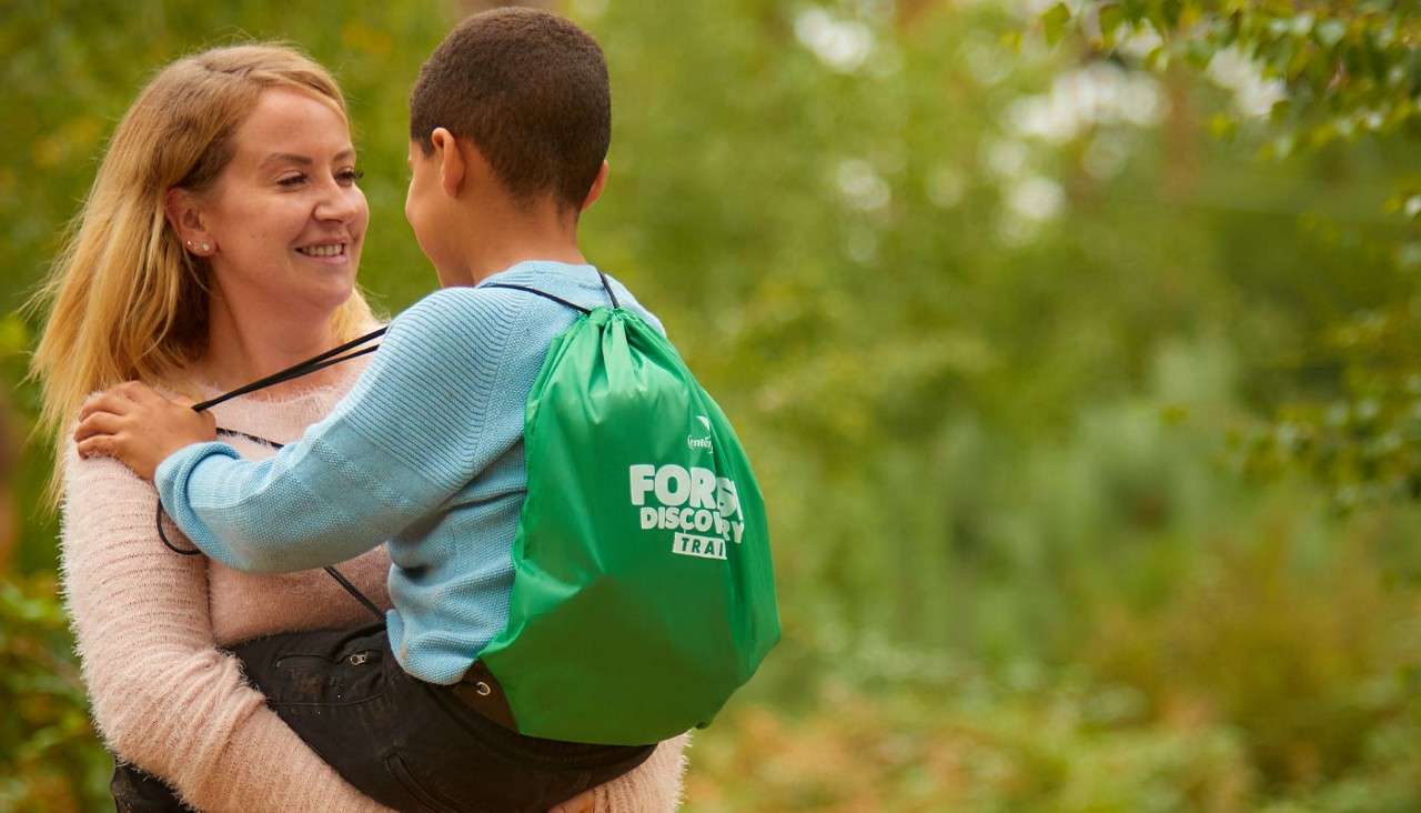 Child with a green drawstring backpack is being carried by an adult, smiling at each other, in a leafy forest setting. Backpack text: FOREST DISCOVERY TRAIL.