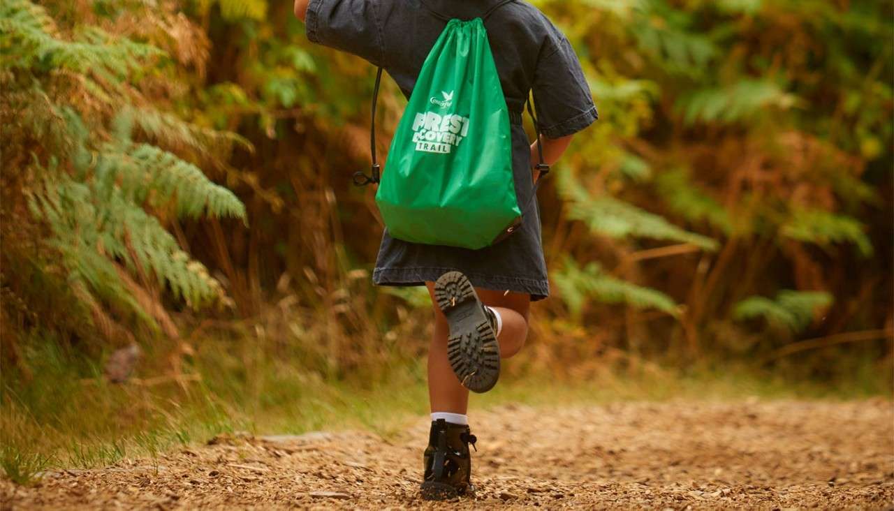 Child with green drawstring backpack runs along a leaf-strewn forest path, surrounded by ferns and warm light. Text on bag: “FOREST DISCOVERY TRAIL.”