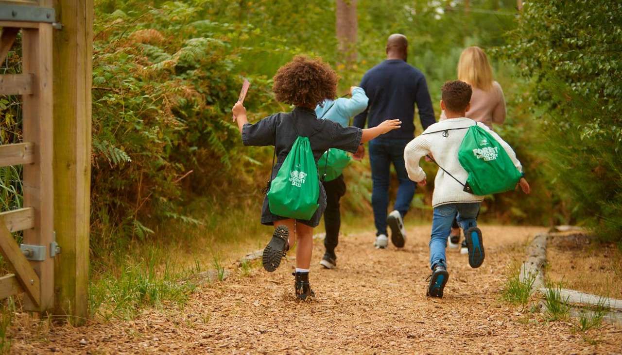 Three children run along a woodchip path, wearing green drawstring backpacks; two adults walk ahead among trees and ferns. Text on bags: “FOREST DISCOVERY [illegible]”.