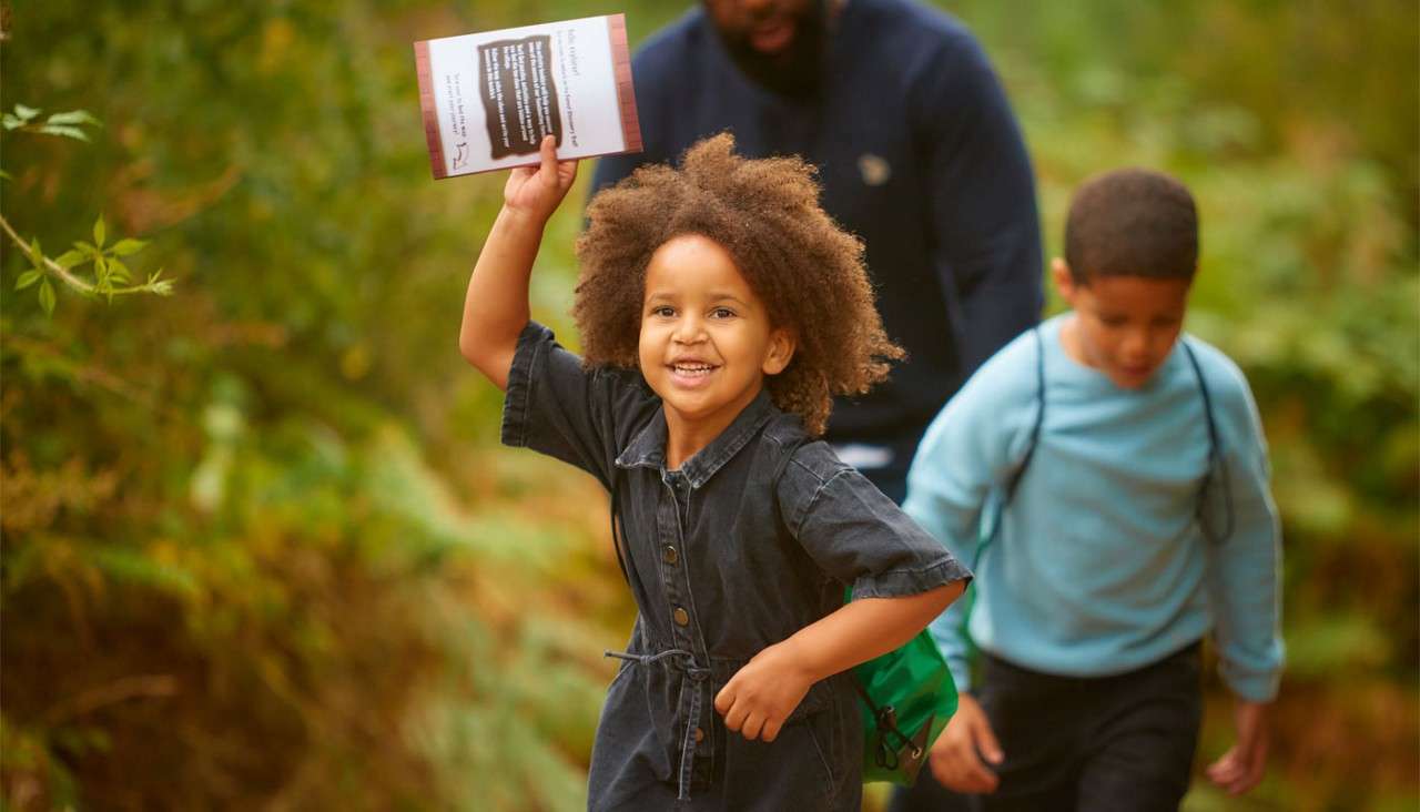Smiling child raises a pamphlet while hiking, leading along a leafy forest path; another child and an adult follow behind.