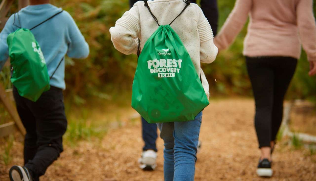 Children wearing green drawstring backpacks walk along a woodland path with adults ahead, exploring a forest trail. Text on bag: “Center Parcs FOREST DISCOVERY TRAIL.”