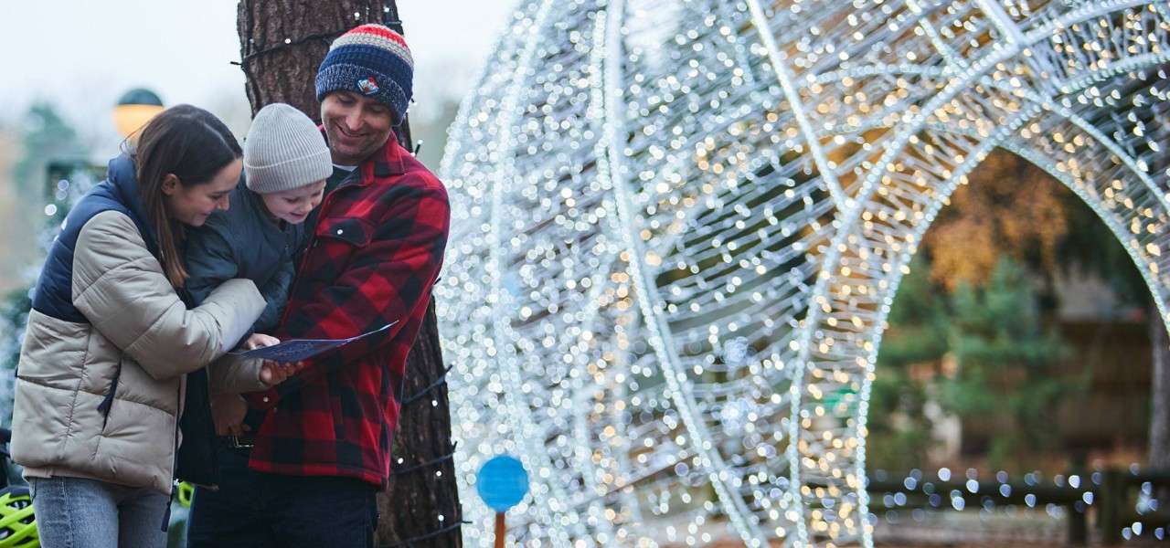 Two adults cradle a bundled child, reading a brochure together, while standing next to a tree and a giant arch of twinkling holiday lights in an outdoor winter display.