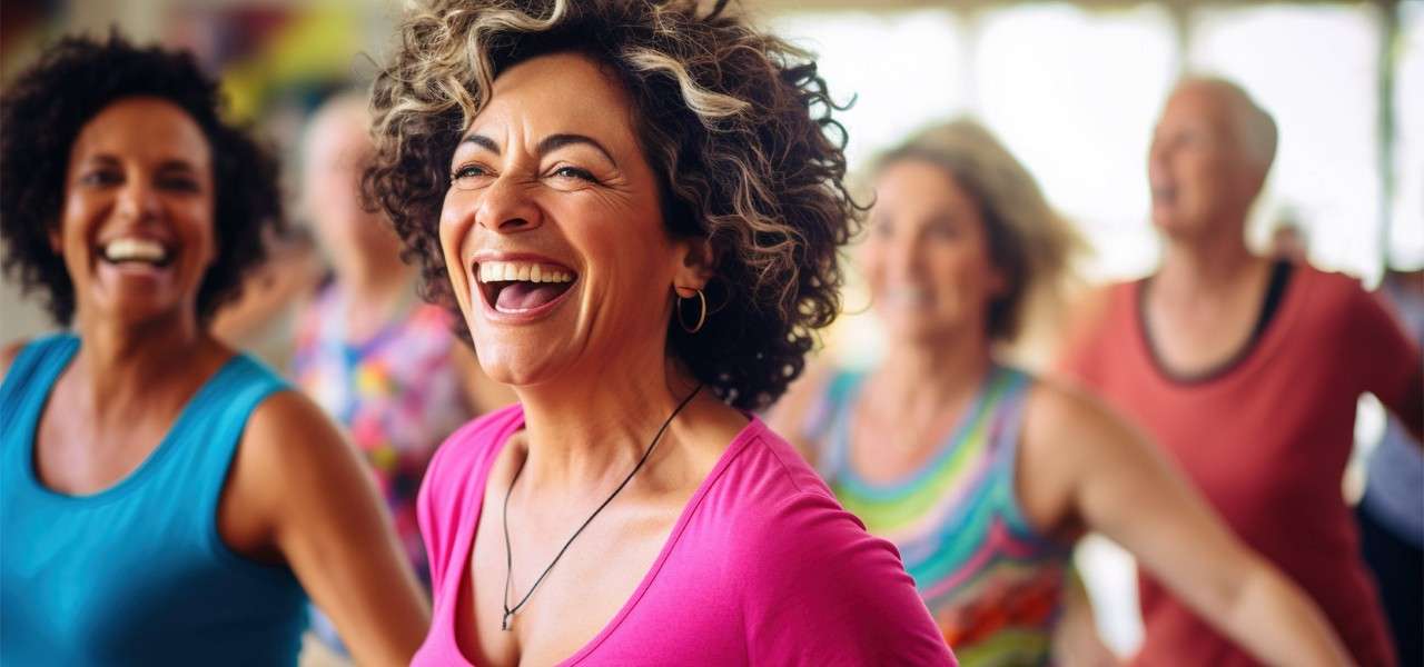 Smiling woman in a pink top laughs mid-dance, moving with others. Background shows several people exercising joyfully in a bright, sunlit indoor studio, wearing colorful athletic clothing.