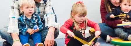 Children play percussion instruments while seated on a carpet, guided by nearby adults; one child shakes a maraca, another taps drumsticks, in a casual indoor group activity.