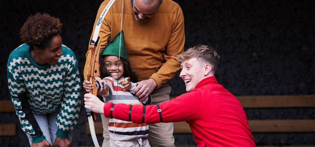 Child archer draws a bow, smiling, assisted by two adults and watched by another, in a covered archery range with benches, creating a supportive, playful learning moment.