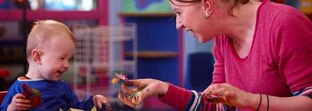 Child and adult finger-paint, laughing and showing paint-covered hands, in a colorful playroom with shelves and toys in the background.