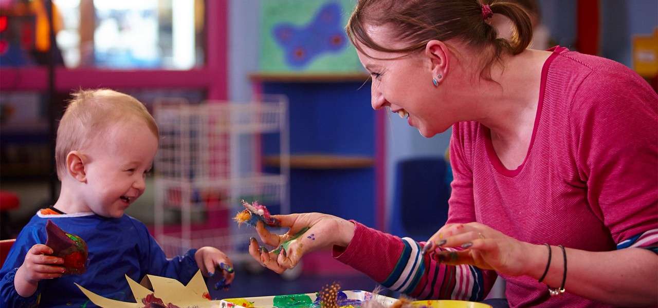 Child and caregiver paint with fingers, laughing together, spreading colorful paint on paper crowns and objects; context: bright classroom with art supplies, pink and blue walls, shelves, butterfly artwork.