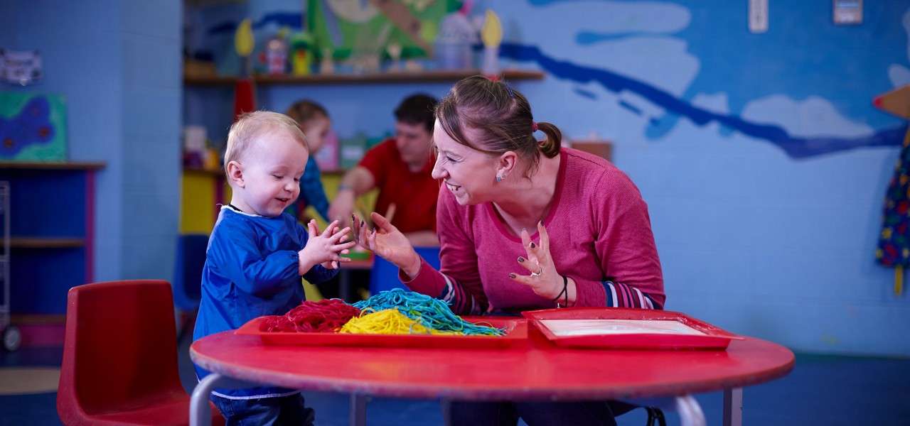 Toddler claps hands while playing with colorful string in a tray; nearby adult smiles and gestures encouragement, seated at a red table in a classroom with murals and other people.
