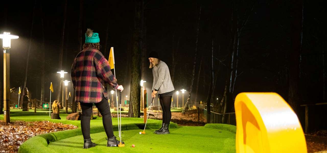 Golfer putts an orange ball toward a hole while another player watches, holding a flagstick; setting: a lit outdoor mini-golf course in a wooded area at night with fallen leaves.