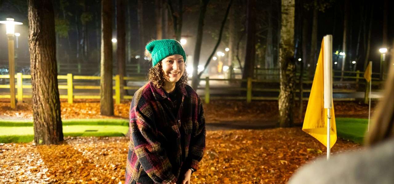 Person in a green knit beanie smiles while standing, hands together, on a nighttime miniature golf course, surrounded by wet leaves, trees, a wooden fence, and yellow flags under lamps.