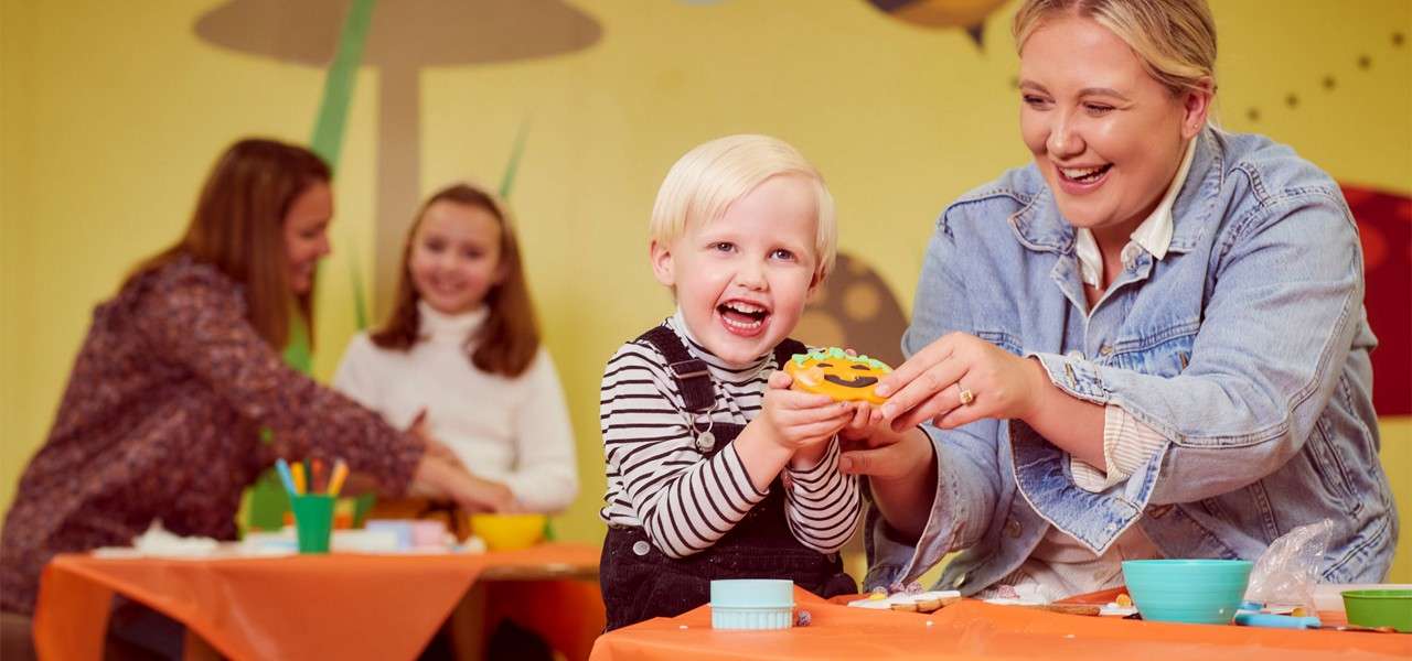 Child decorates a cookie while laughing, assisted by an adult; in the background, two people do crafts at another table in a playful room with bowls, tools, and orange tablecovers.