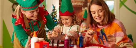 Child paints wooden reindeer with two adults, crafting at a table; elf hats, glue, and colorful paints nearby; festive room with a small lit Christmas tree in the background.