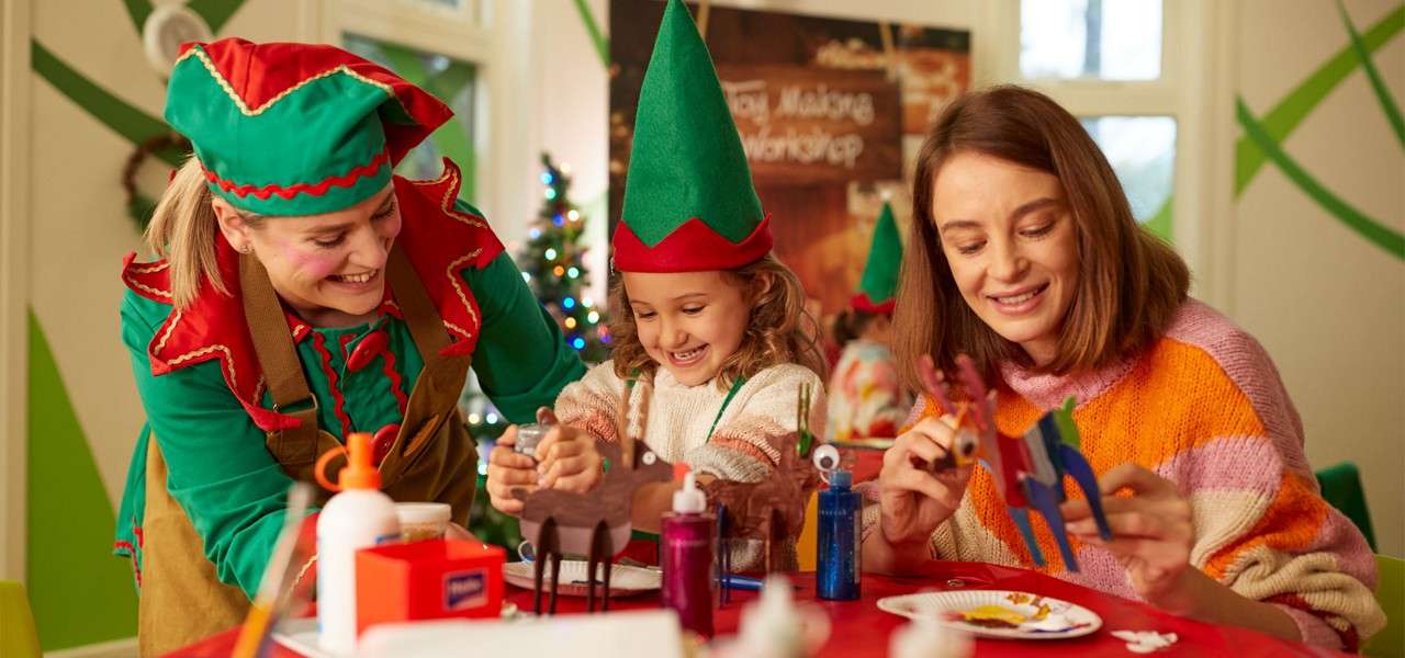 Child in green elf hat glues cardboard reindeer while two adults—one in elf costume—assist; crafting at a festive table with supplies, Christmas tree, and sign. Text: “Toy Making Workshop”.
