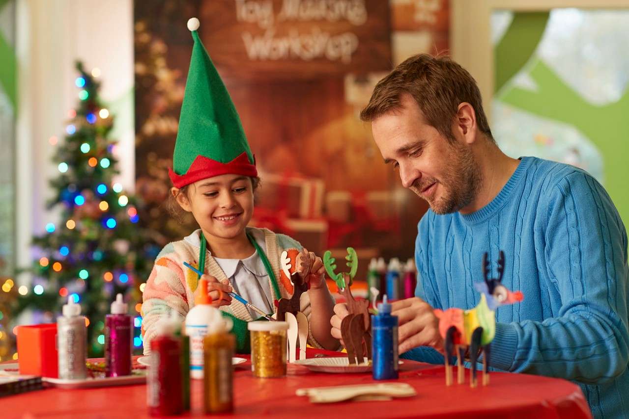 Child in green elf hat glues cardboard reindeer while two adults—one in elf costume—assist; crafting at a festive table with supplies, Christmas tree, and sign. Text: “Toy Making Workshop”.