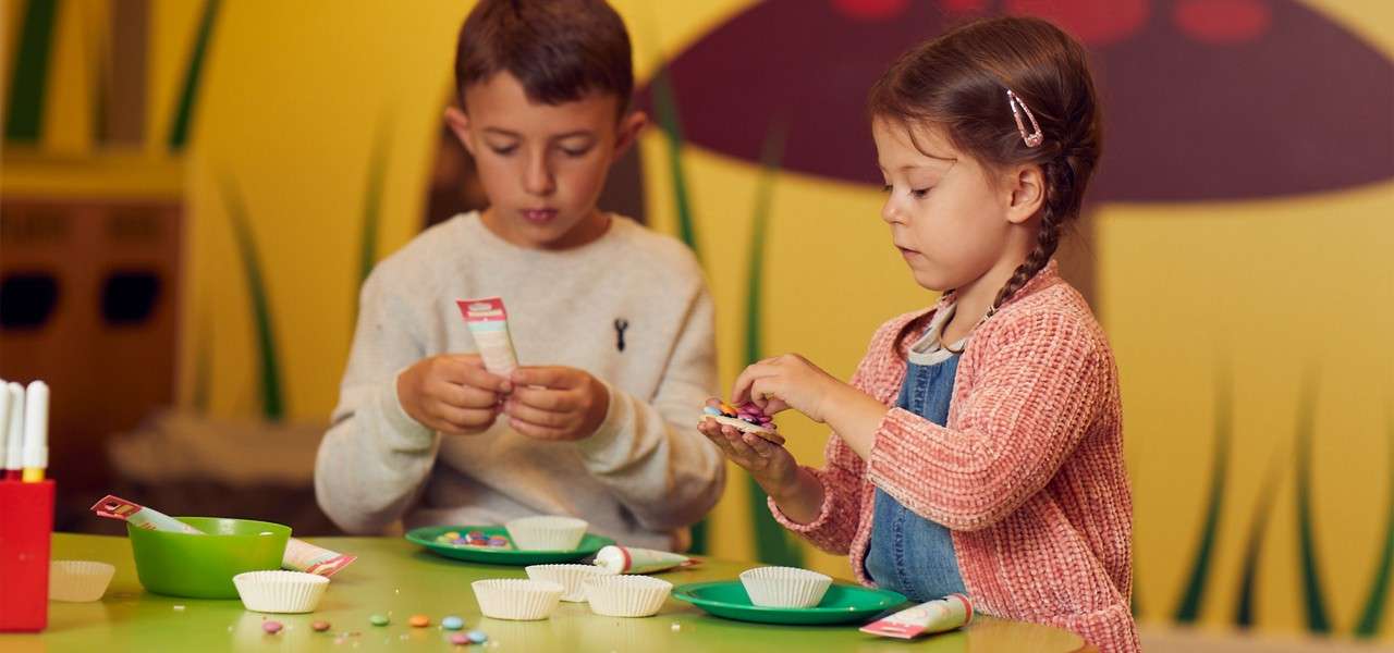 Two children decorate cookies, squeezing icing and placing candy toppings, at a green table with plates, bowls, and paper cups in a colorful, kid-friendly room with grass-like wall art.