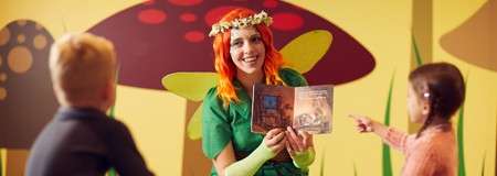 Costumed storyteller reads an open picture book to two children, smiling and gesturing, in a brightly decorated room with oversized mushroom murals and whimsical garden props.