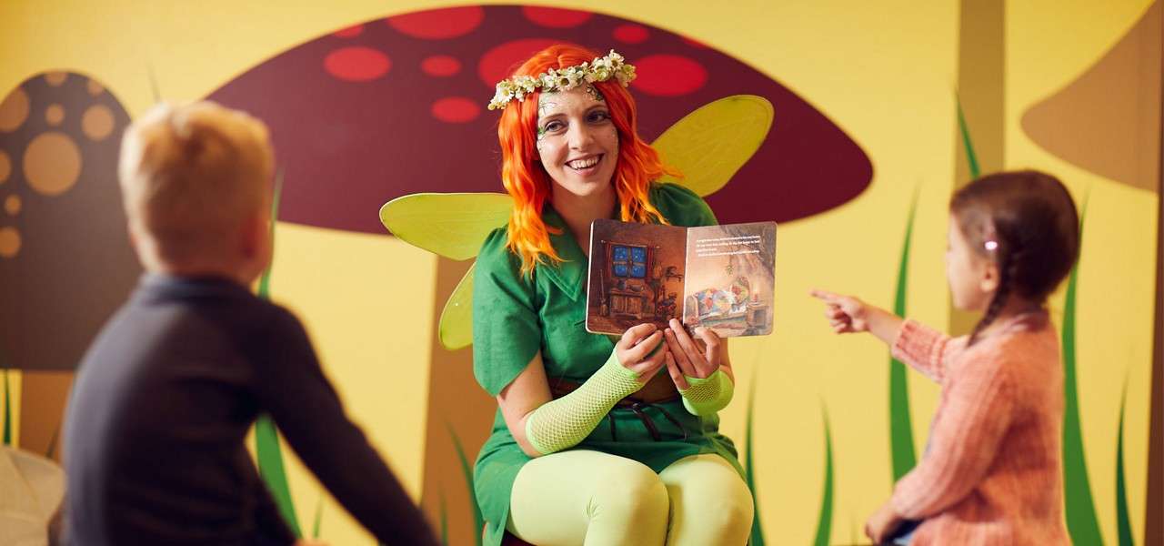 A costumed storyteller in green fairy wings reads aloud from a picture book, engaging two seated children, inside a whimsical mushroom-themed room with bright yellow walls and oversized toadstool graphics.