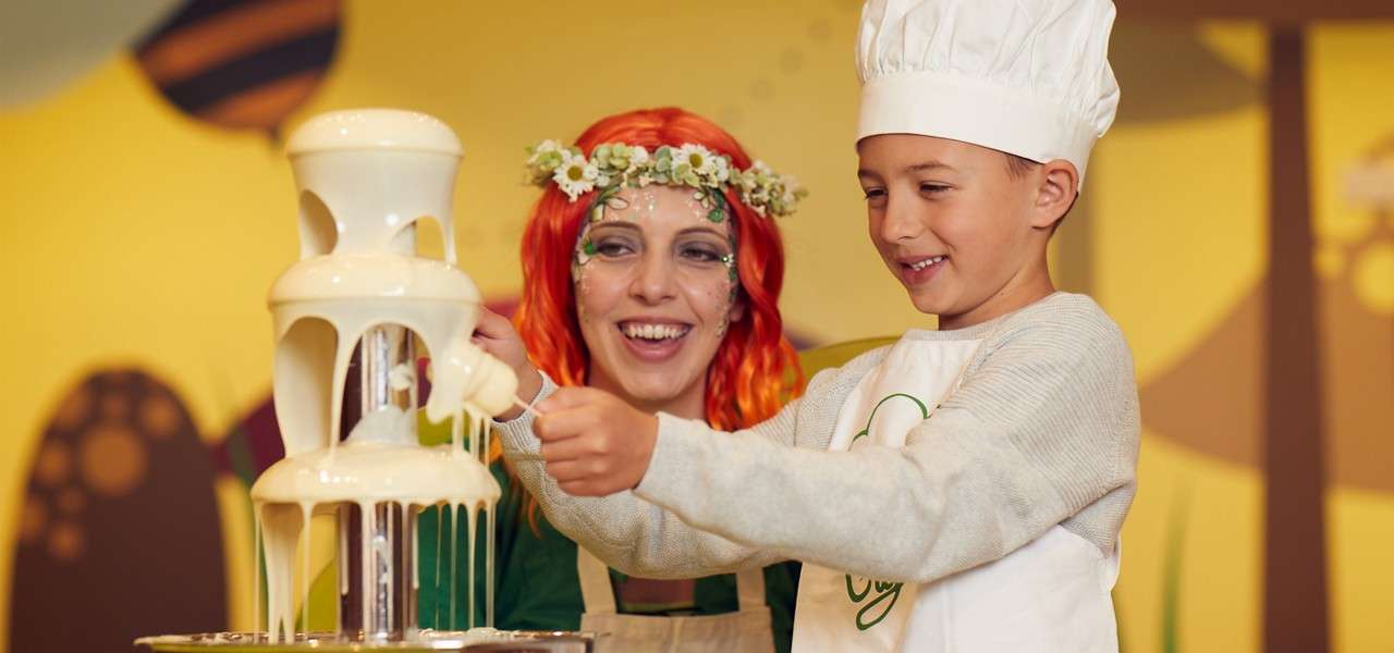 Child chef dips a marshmallow into a flowing white chocolate fountain, guided by a smiling woman with red hair and floral crown, inside a brightly decorated, candy-themed room.