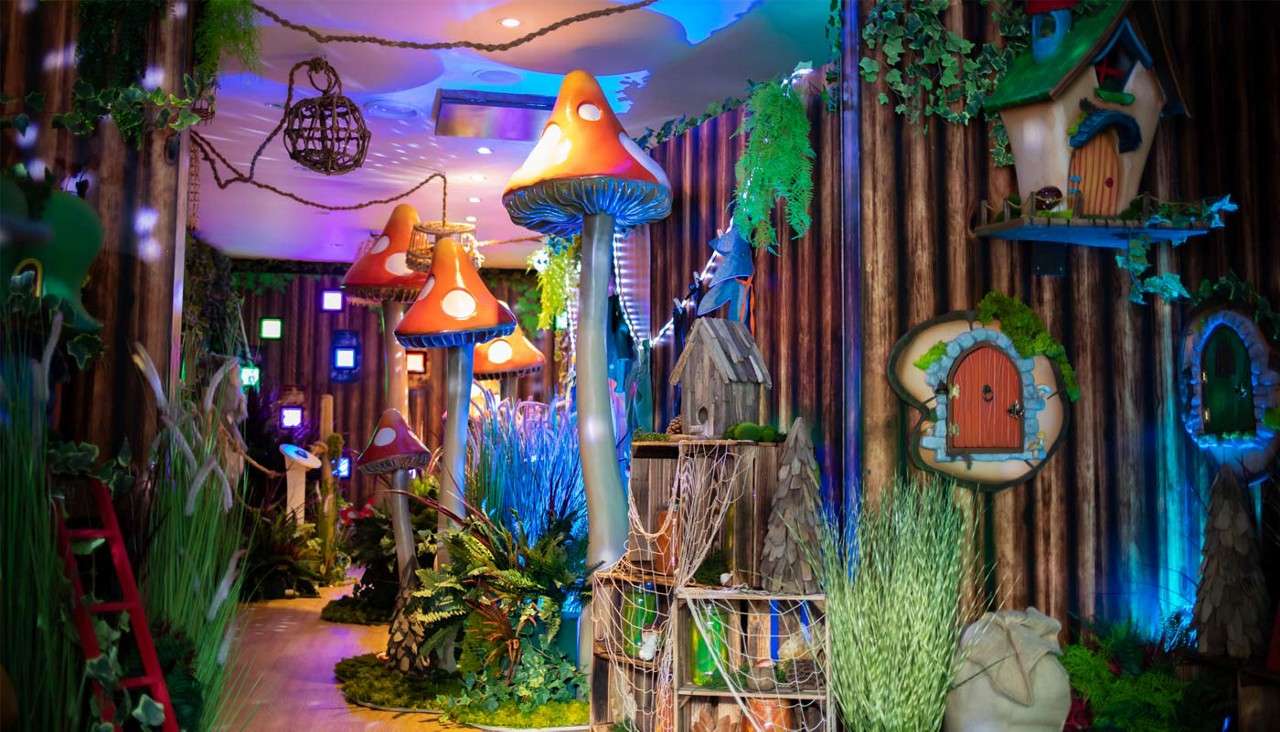 Oversized red-capped mushrooms glow, illuminating a whimsical indoor forest corridor filled with artificial plants, fairy-house doors on log walls, hanging vines, and decorative props.