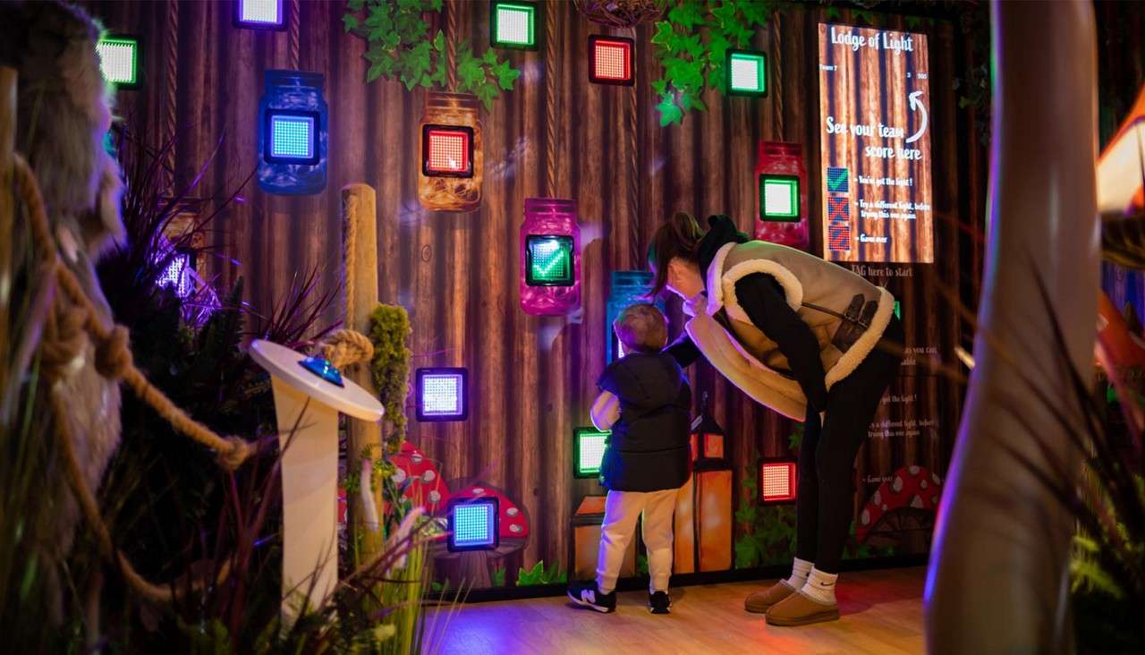 Child and adult tap glowing square buttons; interactive forest-themed wall with jar icons, plants, and mushrooms in an indoor exhibit. Text: “Lodge of Light”; “See your team score here”; “TAG here to Start.”