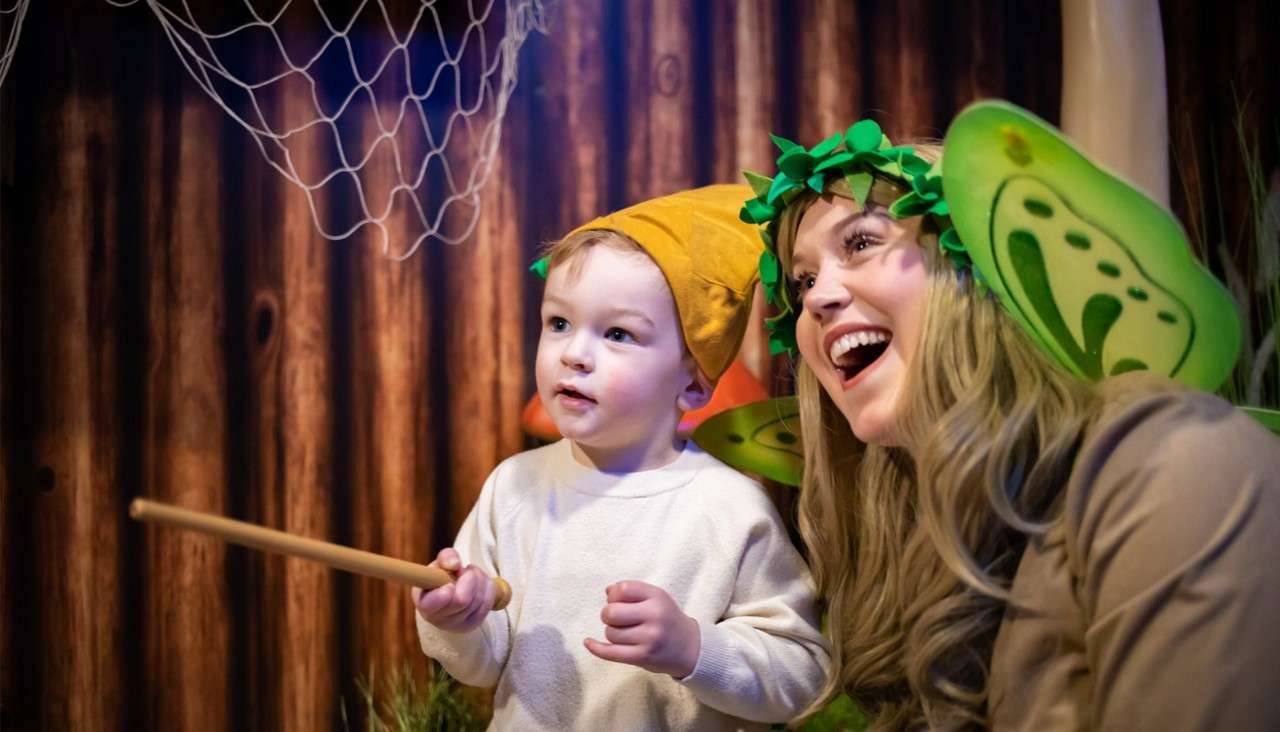 Child points a wooden wand as a smiling woman in fairy wings watches, both in costumes—leaf crown and orange hat—in an indoor, forest-themed setting with netting and wood backdrop.