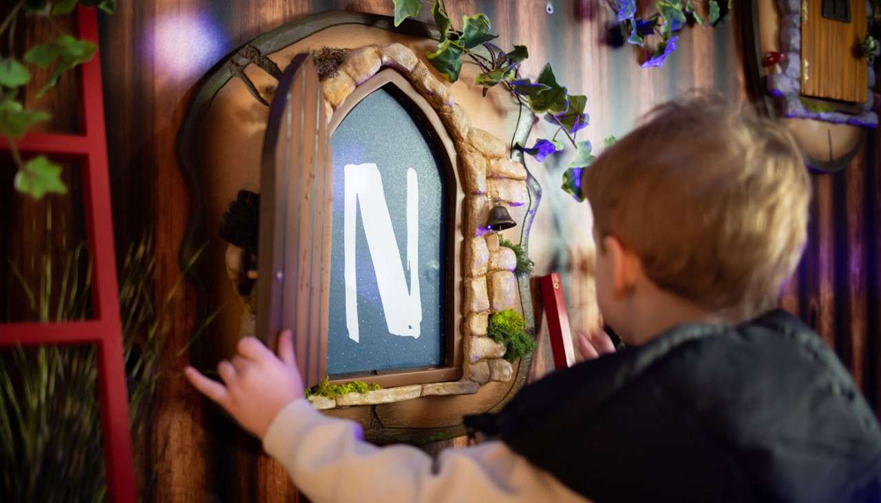 Child opens a small arched wooden shutter, revealing a screen displaying the letter N, within a whimsical stone-framed nook decorated with ivy, moss, and a tiny bell on a themed indoor wall.