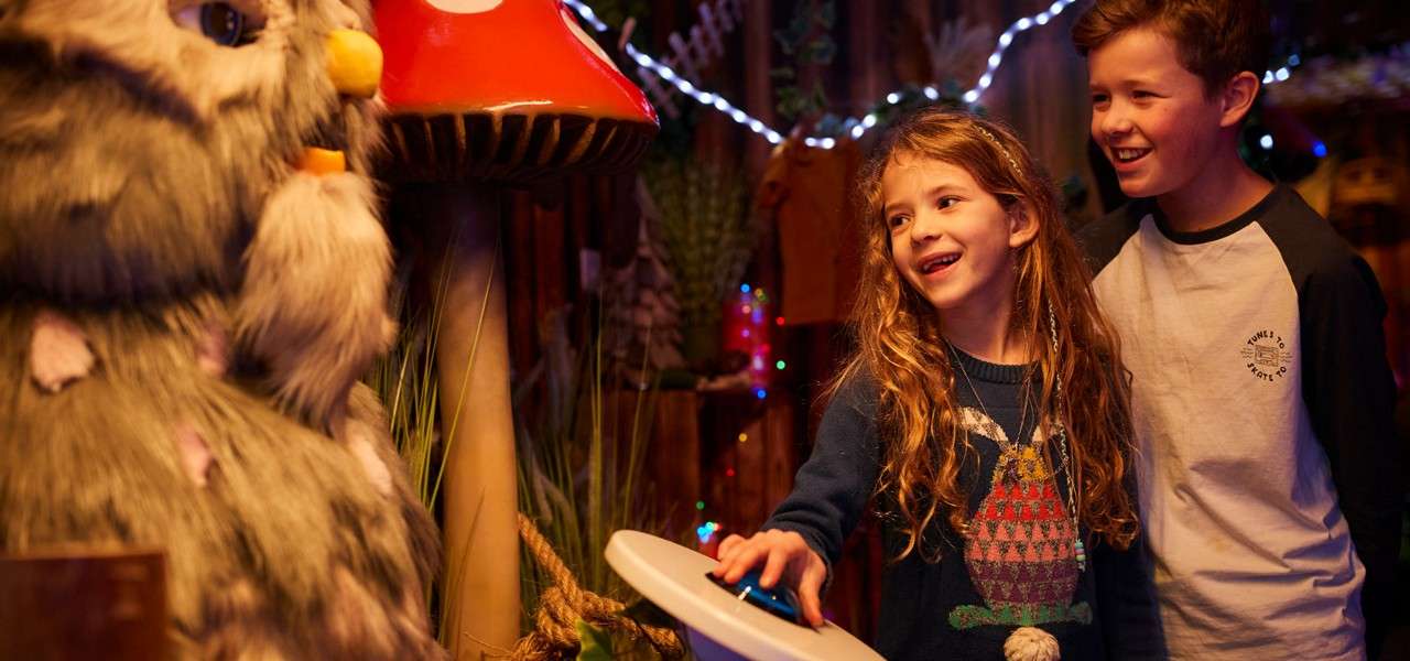 Two children press a glowing button, smiling, while facing a furry animatronic creature; around them, a whimsical indoor set features a giant red mushroom, plants, and festive string lights.