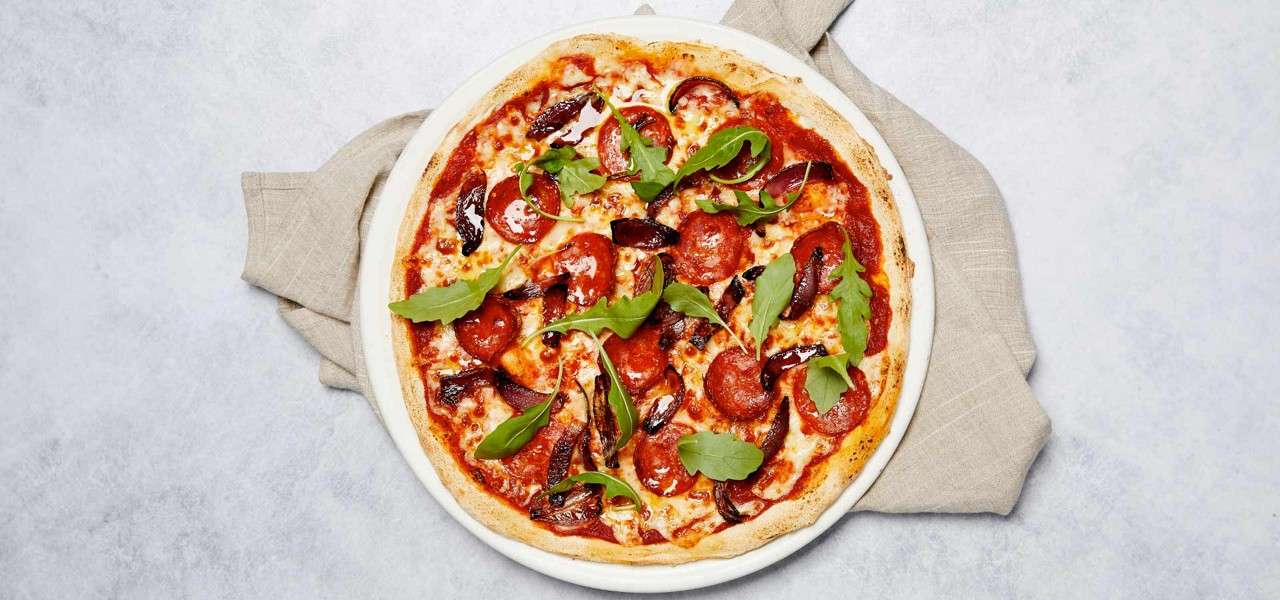 Pepperoni pizza topped with roasted onions and arugula sits whole, cheese melted and glossy, on a white plate placed over a beige napkin on a light stone surface.