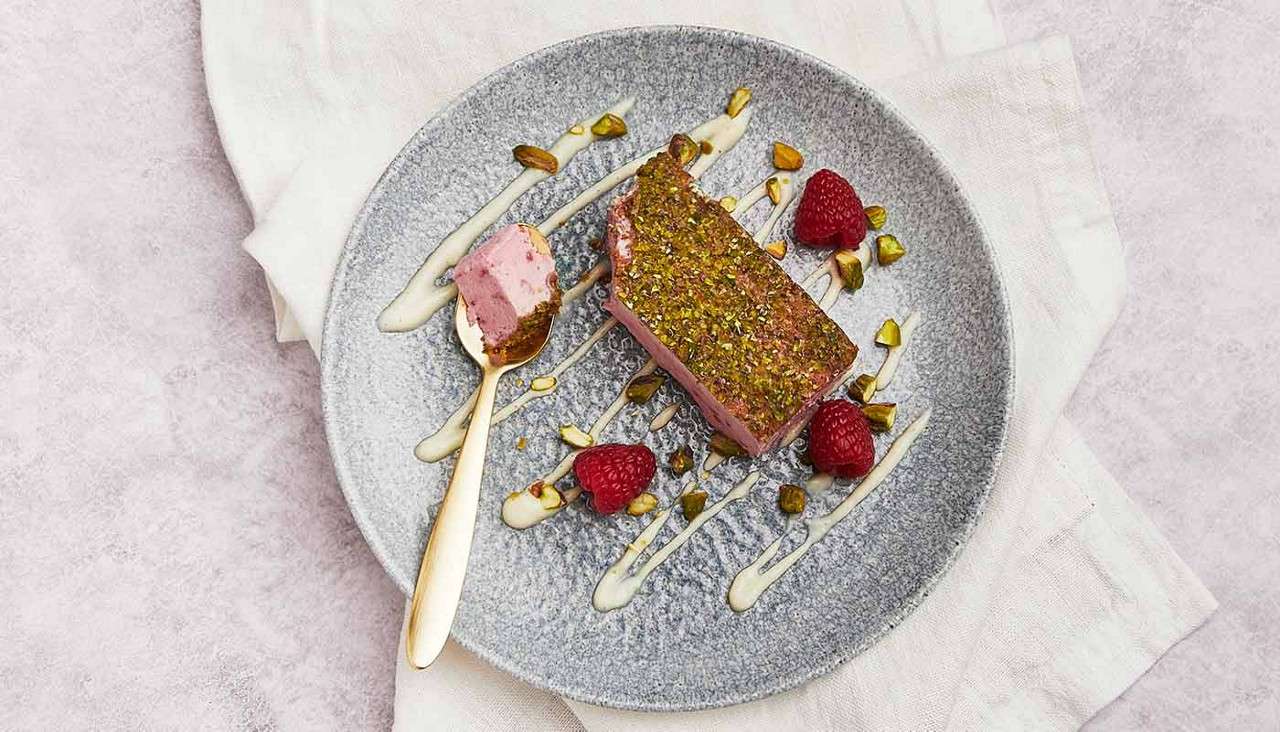 Rectangular pink dessert slice topped with crushed pistachios rests on a gray plate, accompanied by raspberries and cream drizzles; a gold spoon holds a bite. Plate sits on folded linen.
