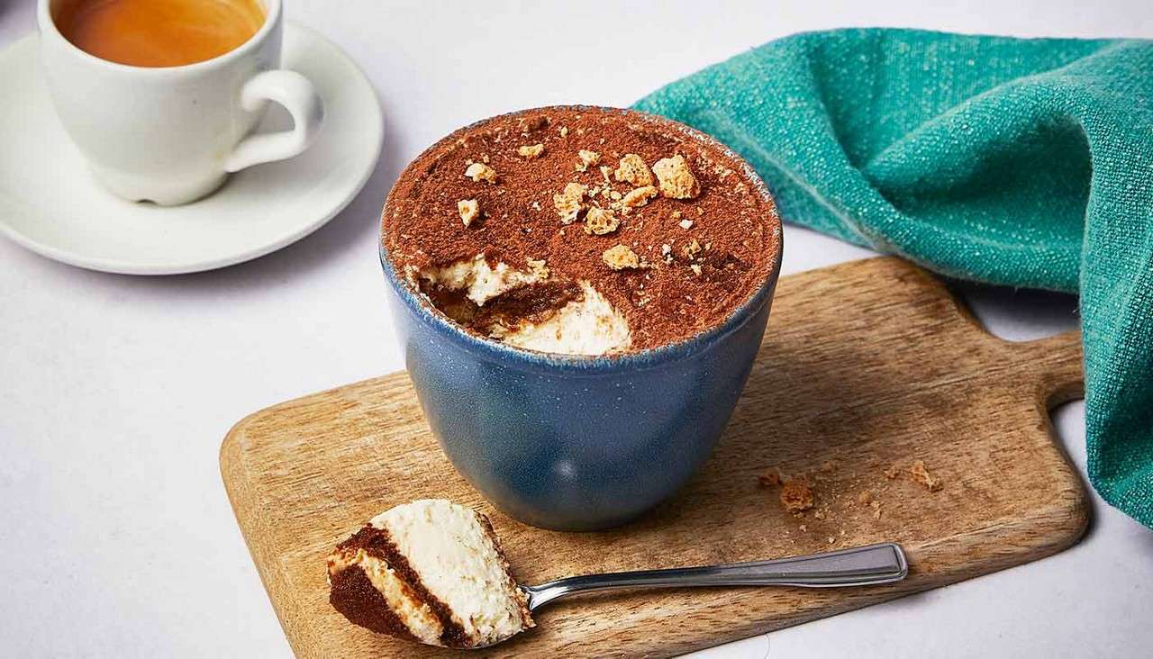 Tiramisu in a blue cup sits with a scoop missing, revealing creamy layers. A spoonful rests on a wooden board, beside a teal napkin and a cup of espresso.