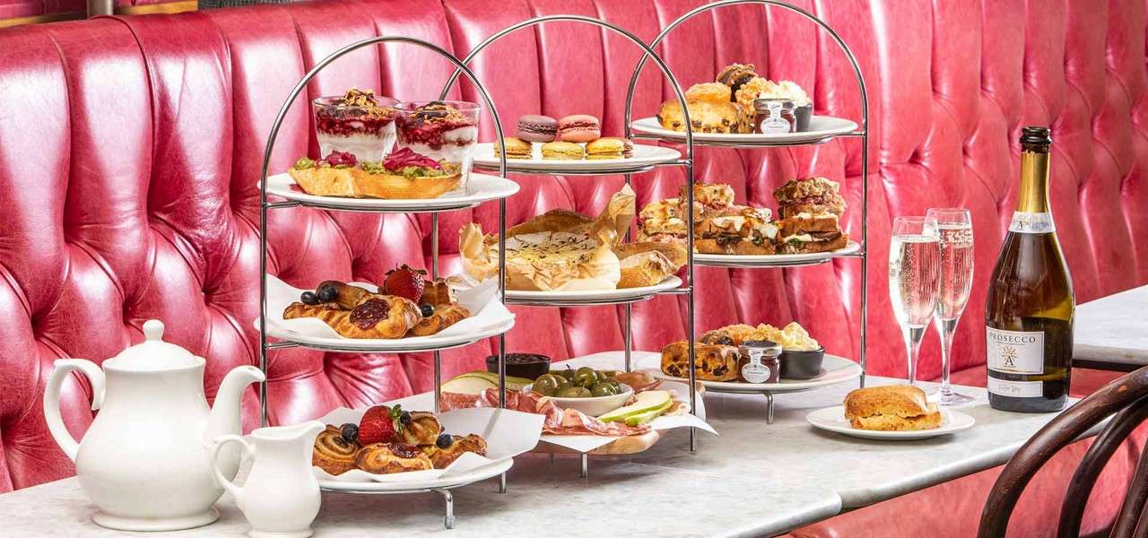 Tiered trays display assorted pastries, sandwiches, and scones; a teapot sits beside olives and macarons, while two flutes stand near a bottle labeled “PROSECCO” on a marble table against a pink booth.