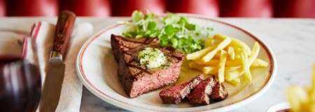 Steak rests sliced with melting herb butter, accompanied by fries and green salad; plate sits on a marble table with knife, napkin, and red booth background, wine glass at side.