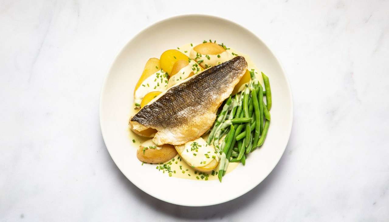 Seared fish fillet lies over sliced potatoes in creamy sauce with chives, accompanied by green beans, arranged neatly on a white plate set on a bright, minimal tabletop.