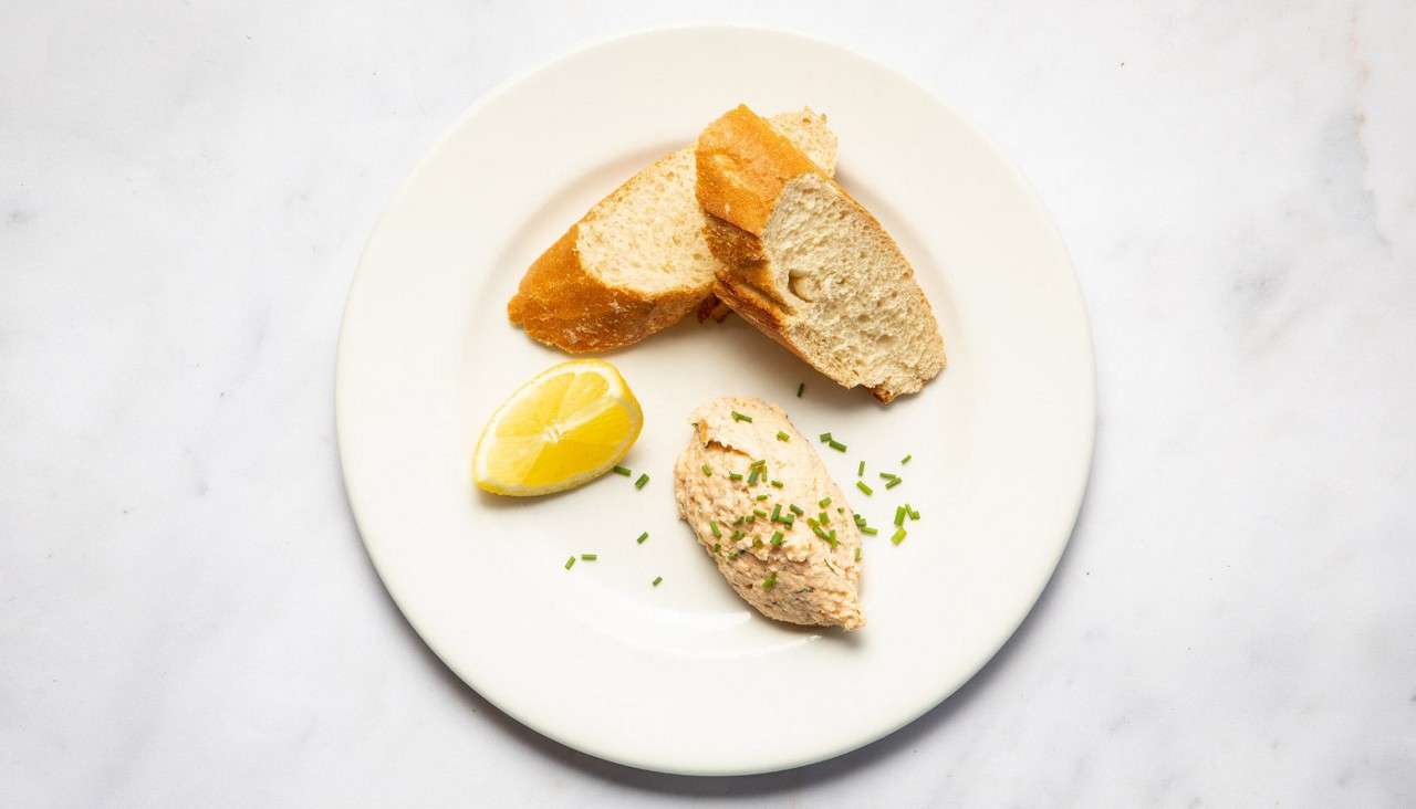 Quenelle of creamy spread sits beside sliced baguette and a lemon wedge, garnished with chopped chives, on a white plate set against a white marble surface.