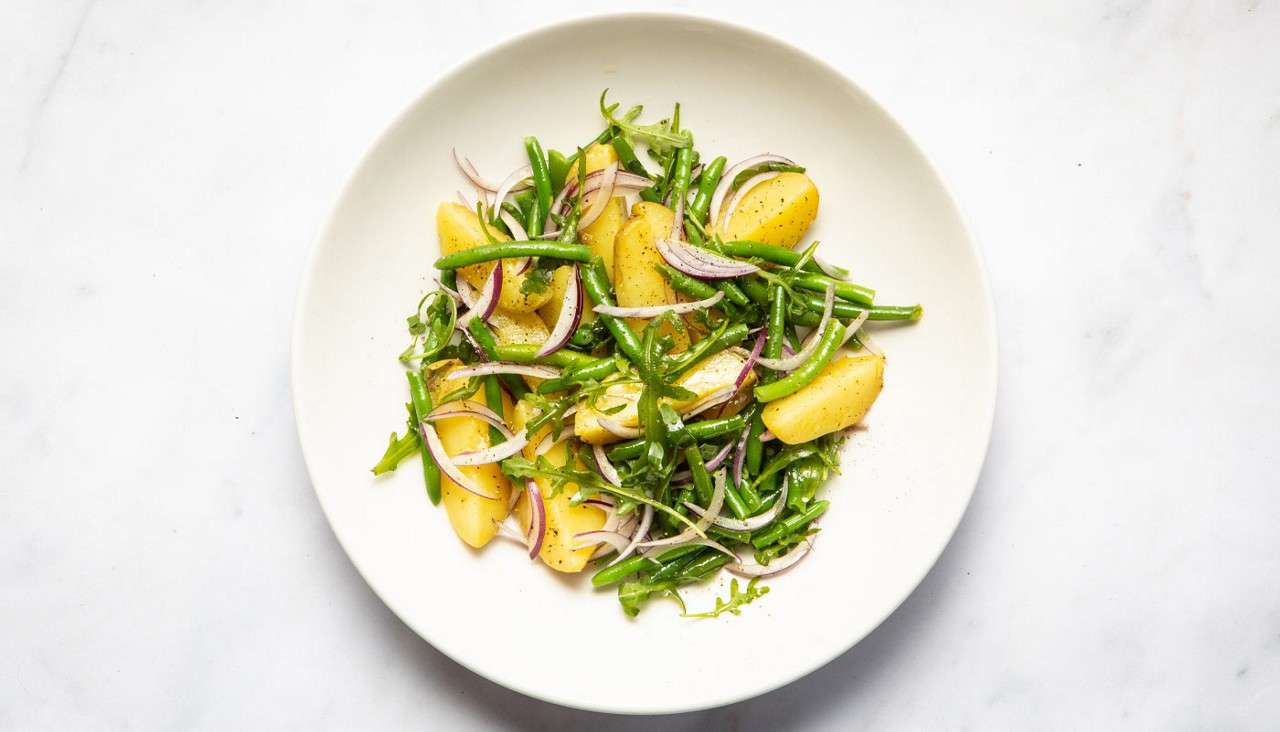Salad sits on a white plate: boiled potato wedges and green beans tossed with arugula, thin red onion, and vinaigrette, sprinkled with pepper, placed on a white marble background.