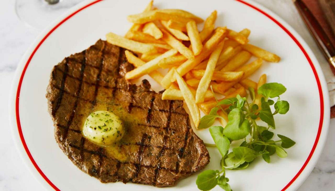 Grilled steak rests topped with melting herb butter, accompanied by golden fries and a small watercress garnish, on a white plate with a red rim set on a marble tabletop.