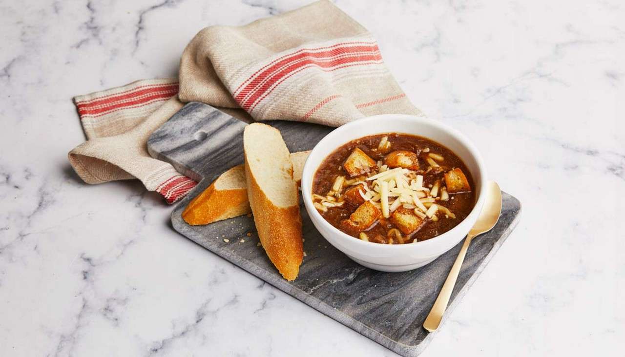 Bowl of dark soup with croutons and shredded cheese sits on a gray board, beside baguette slices and a spoon, with a beige towel on a white marble countertop.