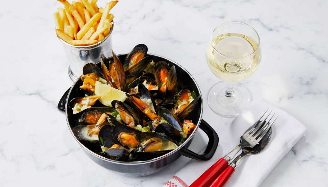 Steamed mussels sit open in a black pot, garnished with lemon and herbs; beside them, a metal cup of fries, a glass of wine, and cutlery on a marble table.