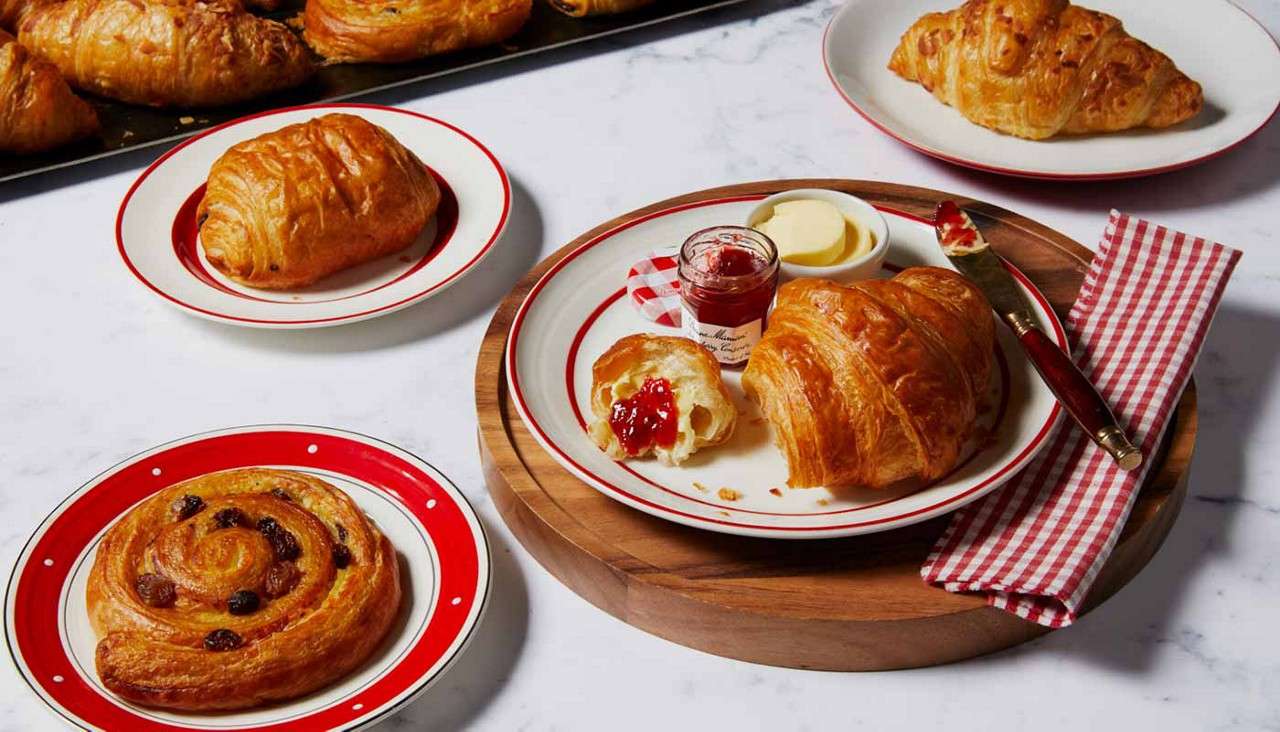 Croissants and pastries sit on red-rimmed plates, one torn open and spread with jam beside a jar and butter. Arranged on a wooden board, marble countertop, checkered napkin nearby.