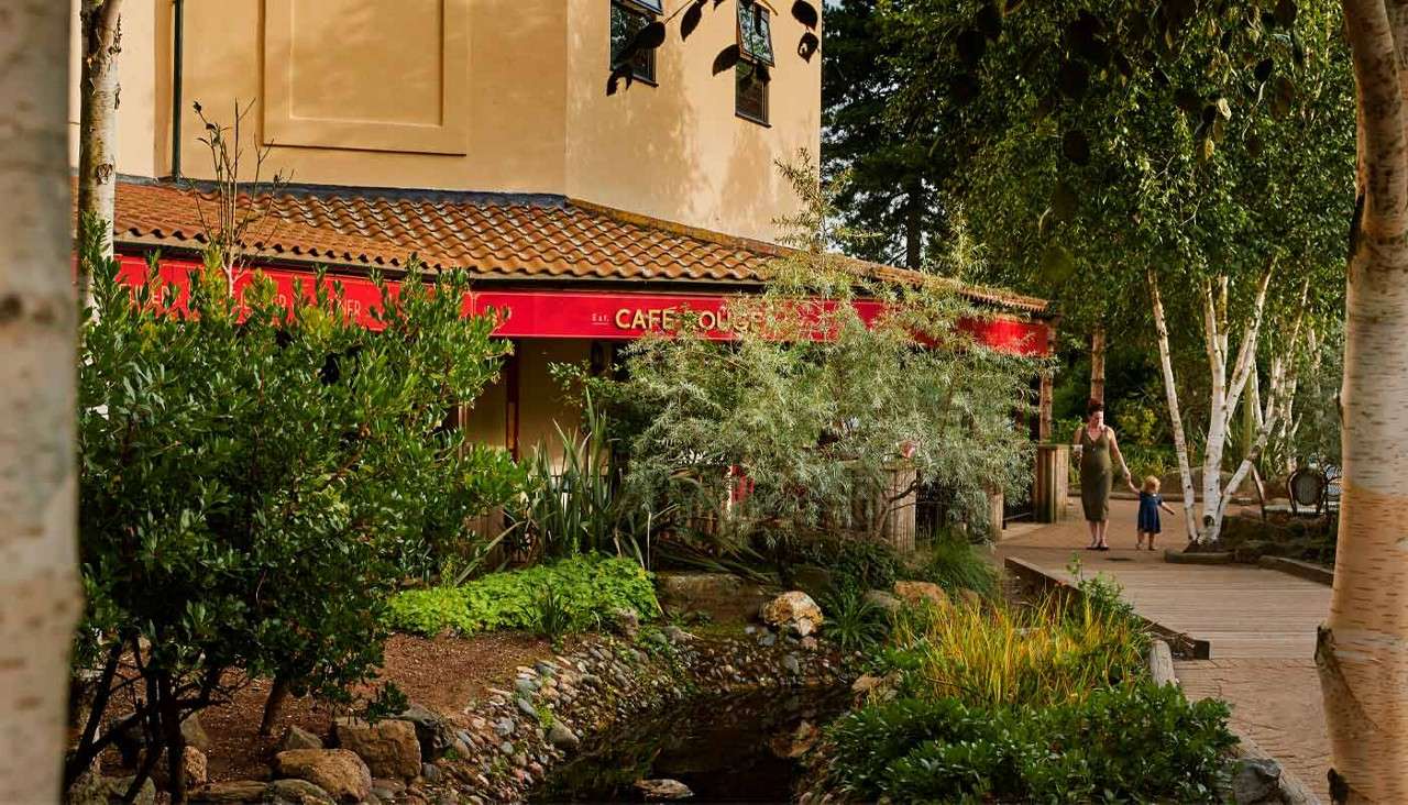 Café entrance—red awning reading “CAFE ROUGE”—rests under trees; a woman holds a child’s hand while walking along a wooden boardwalk beside a small landscaped stream in a garden setting.