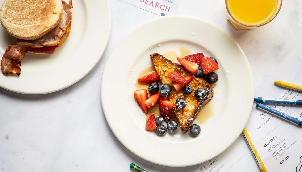 French toast slices topped with strawberries, blueberries, syrup, and powdered sugar sit on a plate, beside a bacon muffin sandwich, orange juice, crayons, and menus showing the headings: starters, mains.