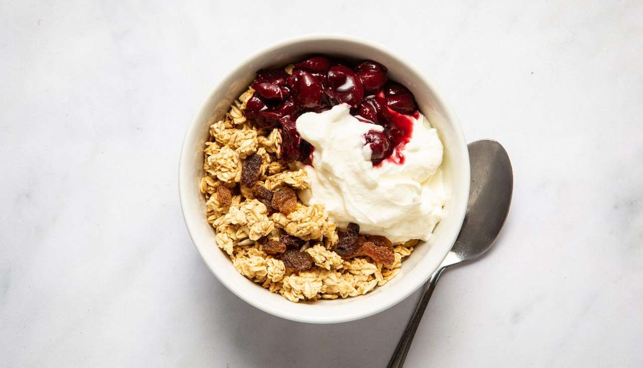 Bowl of granola sits topped with creamy yogurt and glossy cherry compote, raisins mixed in, resting on a white surface with a spoon beside it.