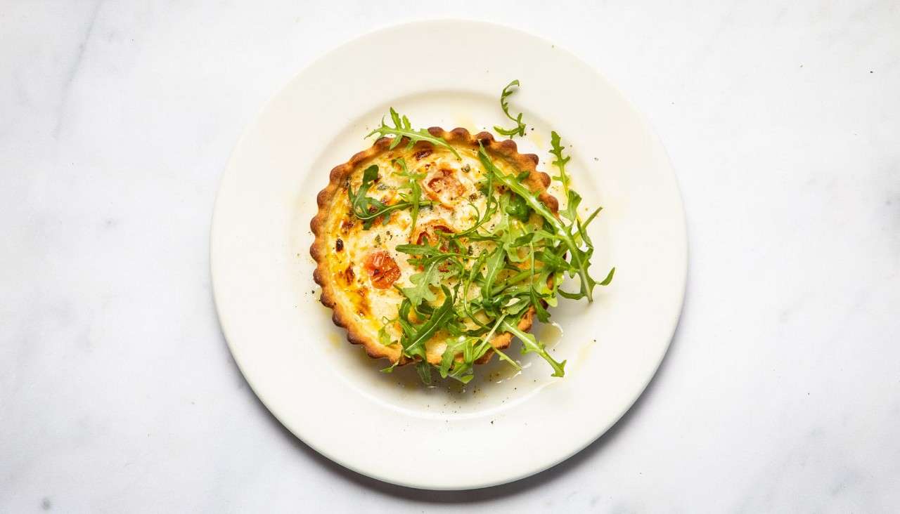 Mini quiche rests on a white plate, topped with scattered arugula, showing browned cheese and cherry tomatoes, set against a clean white marble tabletop.